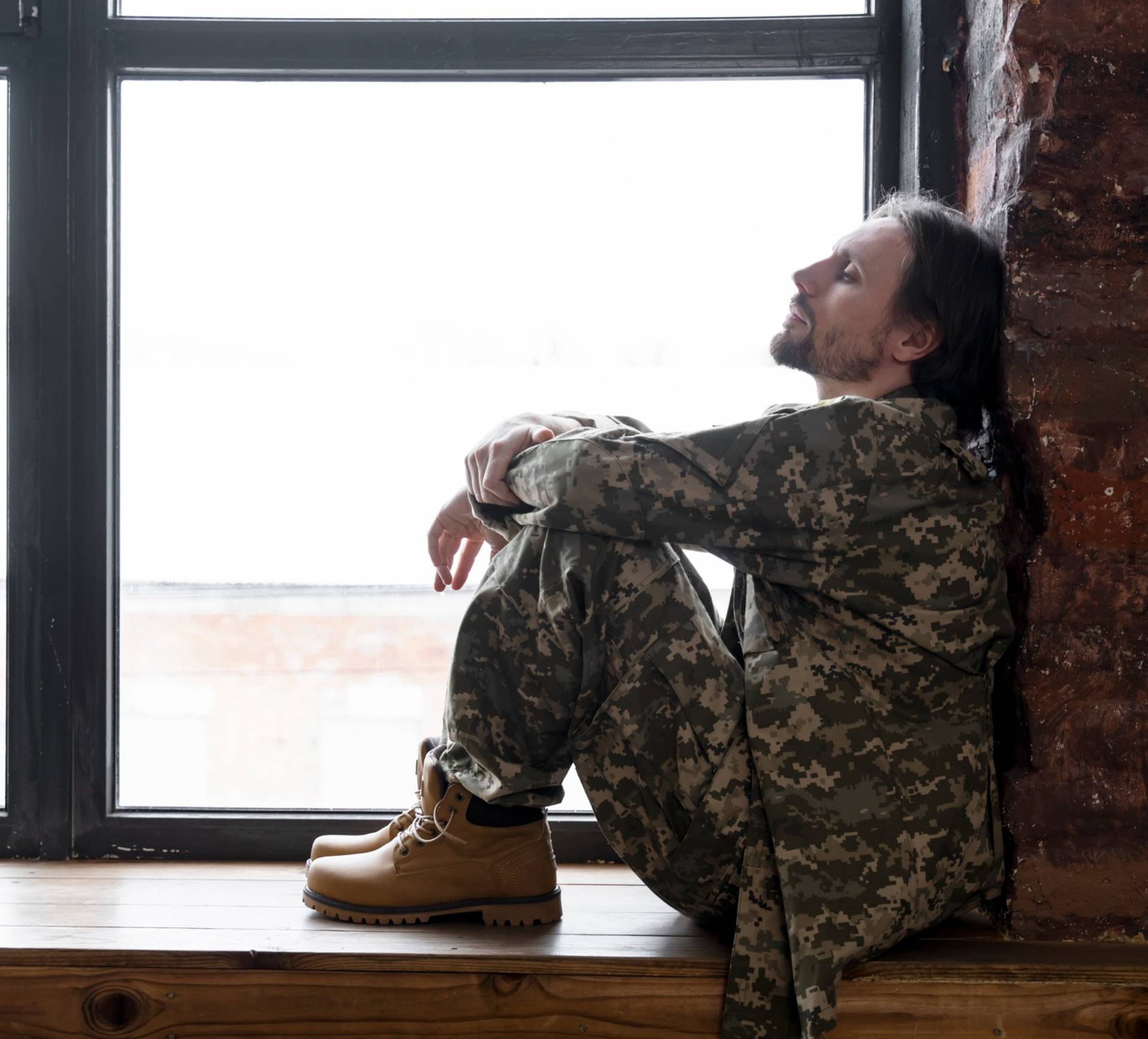 Person in military uniform sitting by a window on a wooden ledge, looking outward.